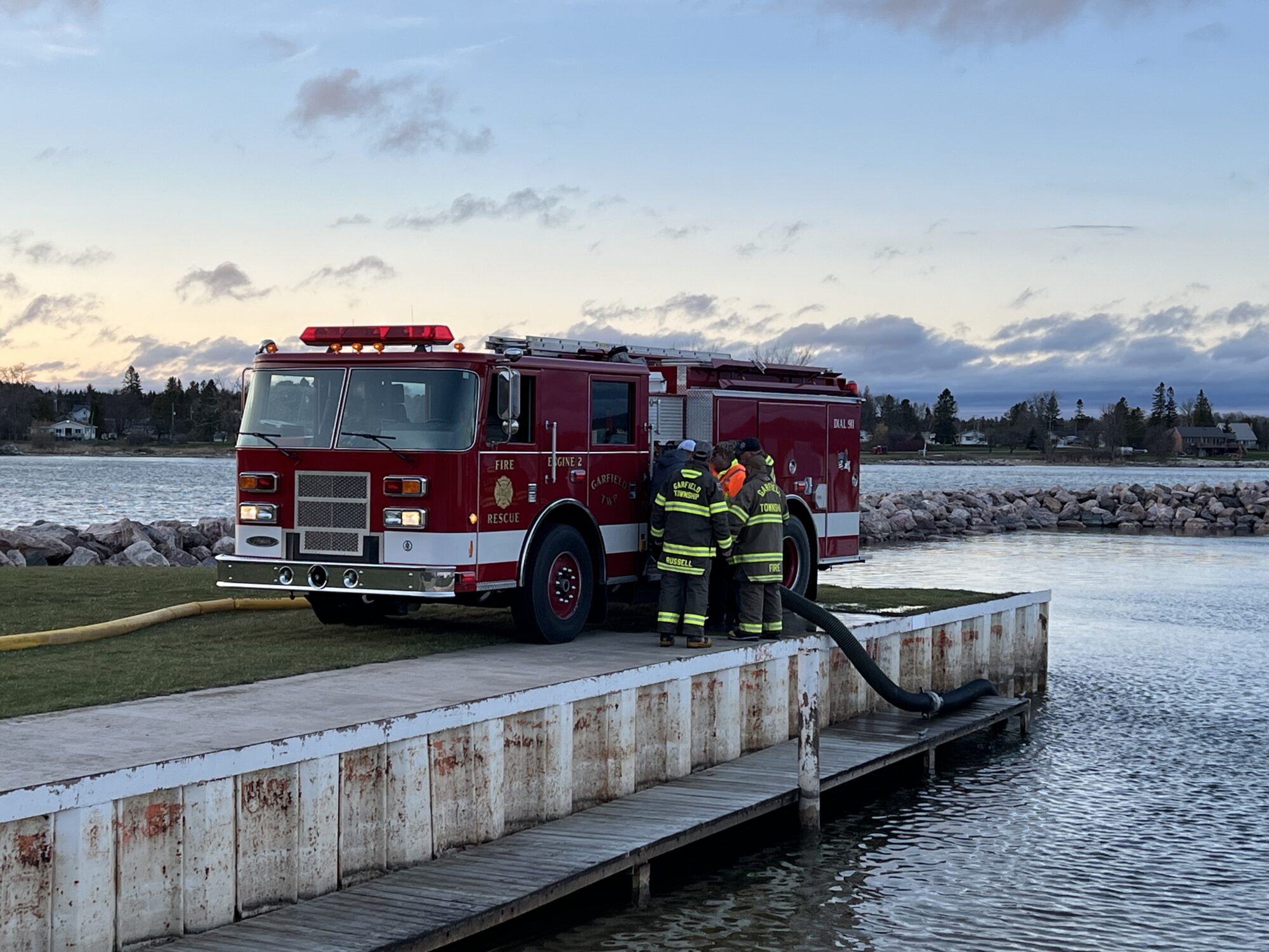 Fire truck near waterfront with firefighters managing hose.